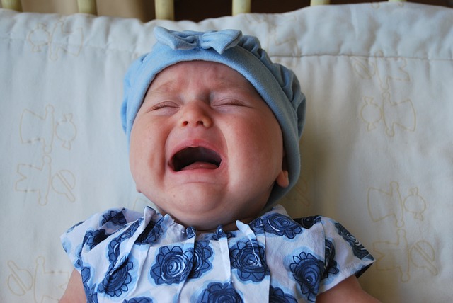 Imagem de uma bebê chorando, usando uma touca azul-claro, enquanto está deitada em uma superfície acolchoada.