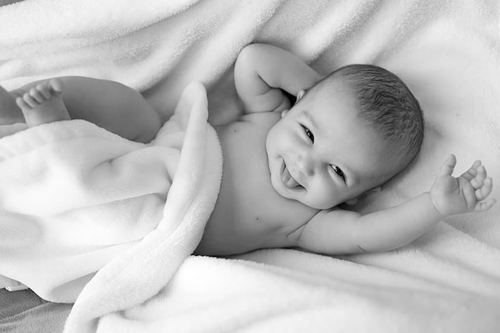  Em imagem preto e branca, um bebê se espreguiça sorridente em meio a um manto/cobertor felpudo.
